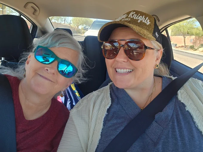 Jackie Harris and Lorea Arostegui on their way to Elko in a car, both wearing seatbelts on a sunny day with trees visible outside