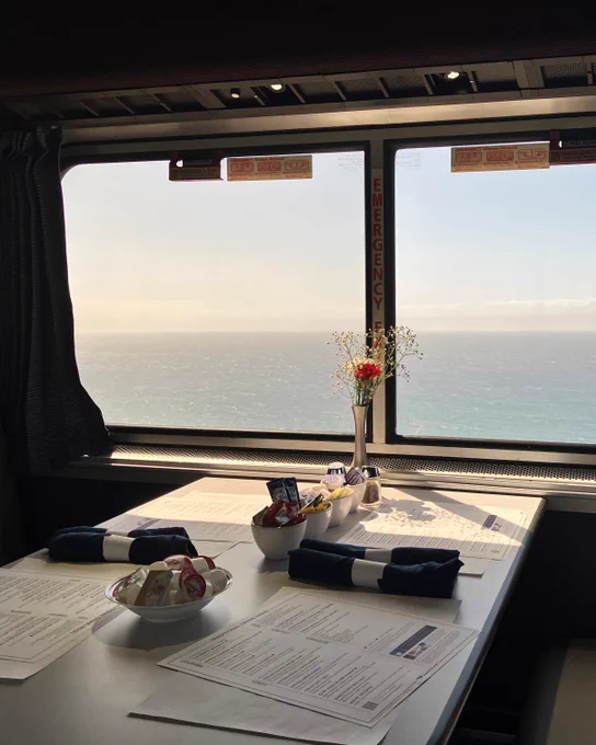 A sunlit table and booth in the Coast Starlight Dining Car, set with menus, bowls of creamer, seasonings and more, and rolled navy napkins. Large windows look out at the ocean and a small vase of flowers sits at the center.