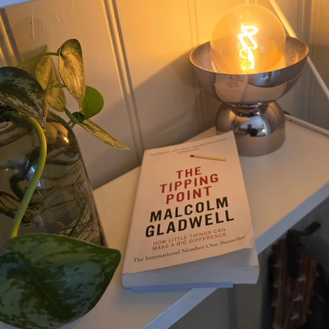 A cozy reading setup with a copy of The Tipping Point by Malcolm Gladwell resting on a white shelf beside a glowing warm lamp and a leafy green plant in a glass vase. Soft golden lighting creates a calm, reflective atmosphere with books partially visible in the background.