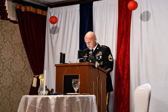A person in a formal military uniform stands at a wooden podium with insignias, delivering a speech. The backdrop features vertical bands of red, white, and blue, with red lanterns hanging. A table with a white cloth, a wine glass, a candle, and a flower is in the foreground.