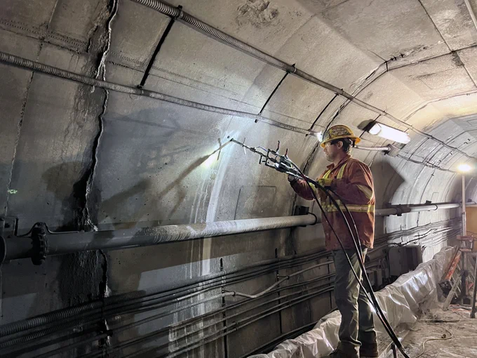 Worker in safety gear using a tool to repair cracks in a concrete tunnel wall illuminated by overhead lights.