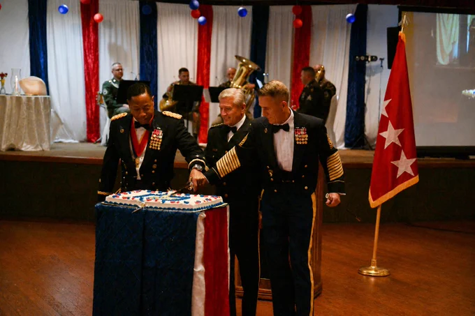 Two people in formal military attire cut a ceremonial cake on a small table. Another person stands beside them. A band plays in the background. Red, white, and blue decorations are visible, along with a flag on a stand.