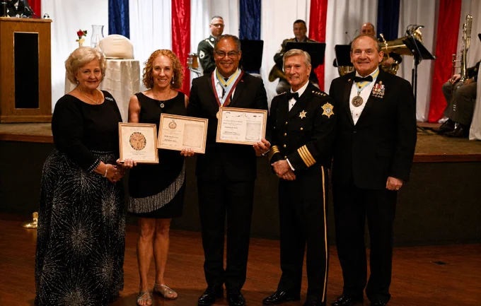 A group of five individuals is standing on a stage at a formal event. Two people are holding certificates, while the others are dressed in military or formal attire. In the background, a band is playing with drums and brass instruments. The setting is decorated with red, white, and blue drapes.