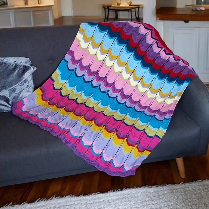 A colourful hand knitted blanket featuring scalloped patterns in hues of blue, pink, yellow, red, and purple is draped over a grey sofa. The room in the background has white cabinetry, a wooden table, and a wooden floor with a light area rug.