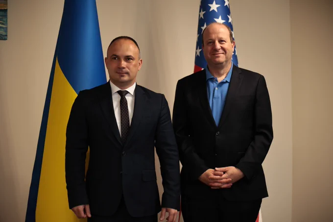 Two men in formal suits stand side by side in front of Ukrainian and American flags indoors.