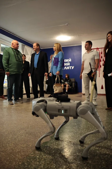 A robotic dog stands on a polished floor while a group of people engage in conversation in the background inside a modern room.