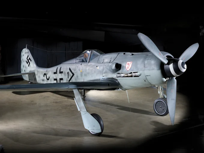 A Focke-Wulf Fw 190, a World War II-era German aircraft, displayed in a dimly lit hangar, featuring its distinctive camouflage and markings.
