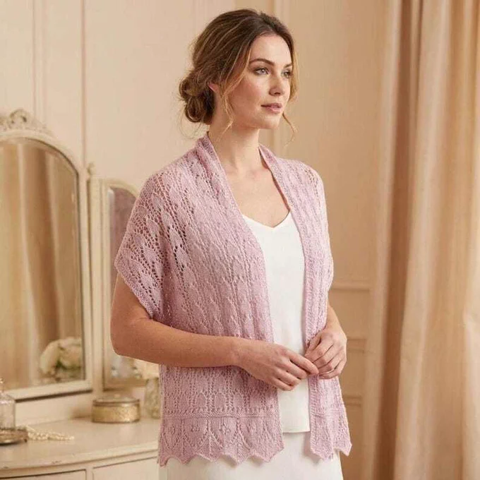 A woman stands wearing a soft pink lace knitted scarf over a simple white top. The scarf features an intricate openwork pattern and a scalloped edge. She is in an elegant room with cream walls, tall draped curtains, and an ornate vintage vanity table with mirrors and decorative items, creating a warm and sophisticated atmosphere.