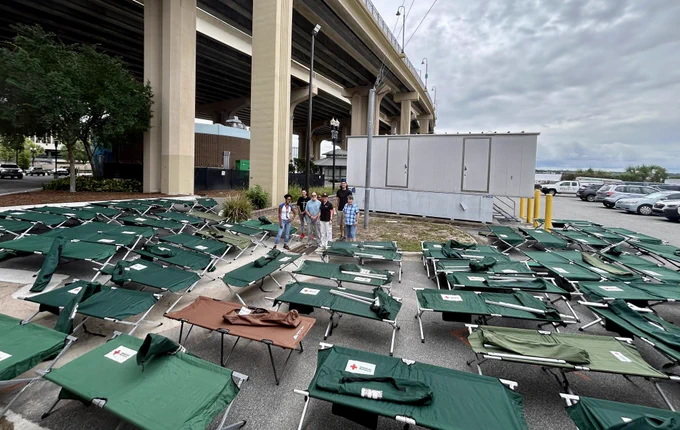 A group of people stands among numerous green cots set up in rows outdoors under a large overpass. The cots display the American Red Cross logo. A small building is visible in the background. The sky is cloudy.