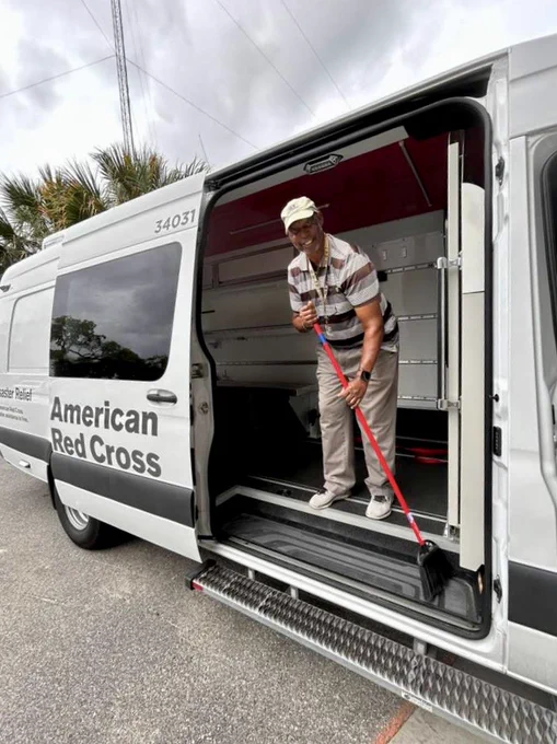 Individual sweeping inside an American Red Cross disaster trailer, smiling. The trailer is parked outdoors on a cloudy day.