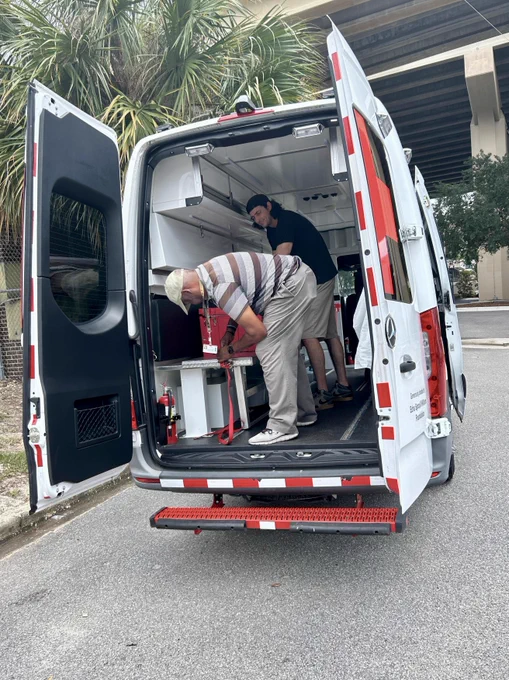 The image shows two people working at the open back of a white van. The van is equipped as a mobile work unit with shelves and equipment. It is parked on the side of a street with some greenery and an overpass in the background.