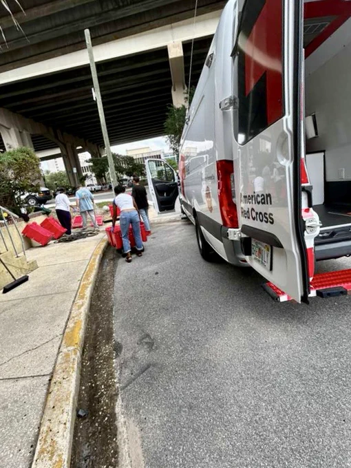 A group of people is unloading supplies from an American Red Cross vehicle parked on the street under a large overpass. The vehicle's large side door is open, and several red containers are on the ground nearby. Trees and urban infrastructure are in the background.