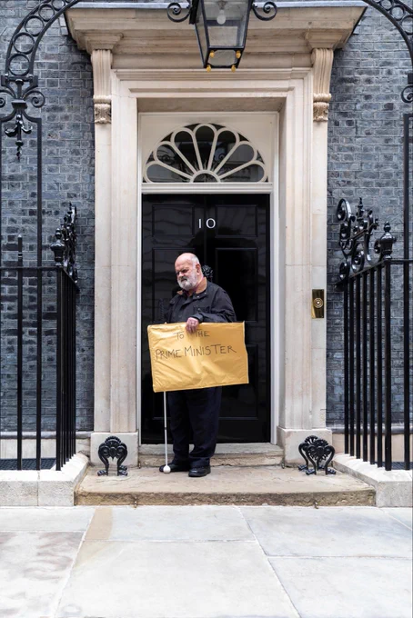 Kevin holding a petition envelope with the words ‘To the Prime Minister’ outside 10 Downing Street, holding his white cane.