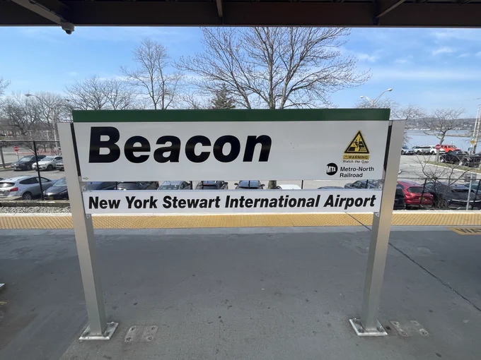 A sign at a train station platform displays "Beacon" and "New York Stewart International Airport" with the Metro-North Railroad logo. Cars and leafless trees are visible in the background.
