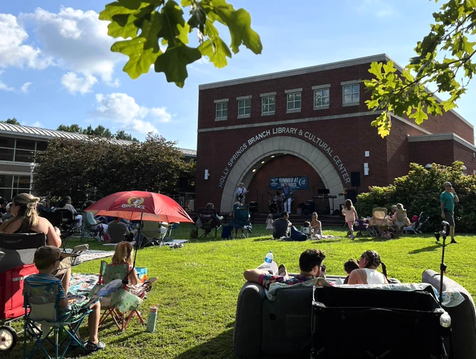 A crowd of people is gathered on a grassy area outside the Holly Springs Branch Library & Cultural Center. Some are sitting on chairs or blankets, while others are in strollers. A red umbrella and trees provide some shade. The library's brick building and entrance arch are visible in the background.