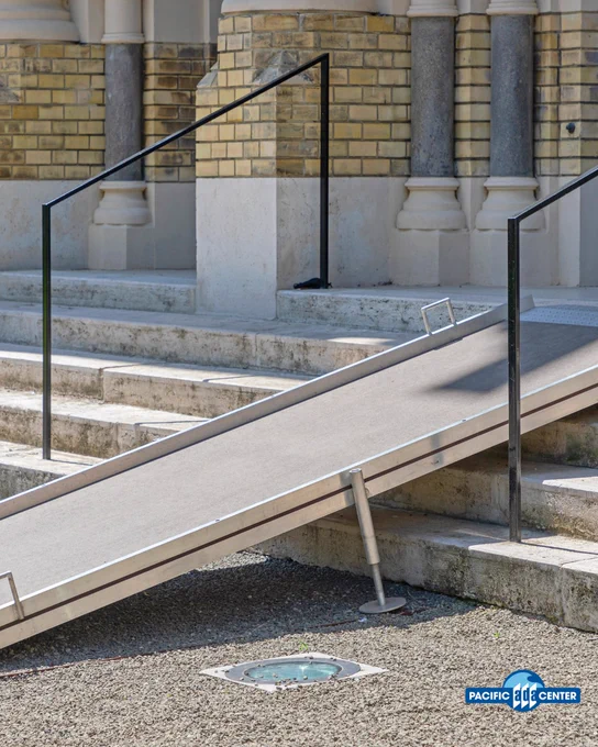 Concrete accessibility ramp with metal handrails leading up to a building entrance with stone steps and brick walls.
