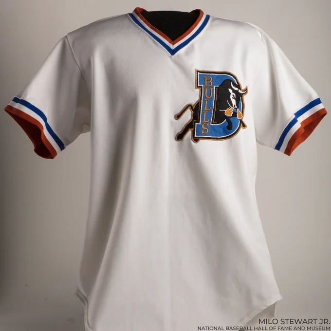 Home white Durham jersey worn by Tim Robbins as he portrayed Calvin "Nuke" LaLoosh. The jersey features a bull charging through a blue block-D logo with blue, white and orange trim around a V-neck cut and sleeves. (Milo Stewart Jr./National Baseball Hall of Fame and Museum)