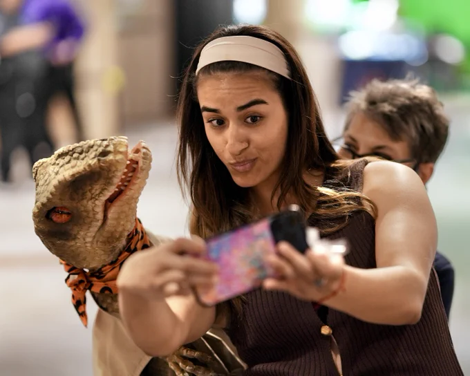 A woman is taking a selfie with a realistic dinosaur puppet wearing an orange bandana in an indoor setting.