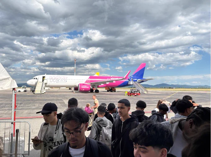 Passengers line up to board a Wizz Air plane on a cloudy day. The vibrant pink and purple aircraft stands out against the gray sky.