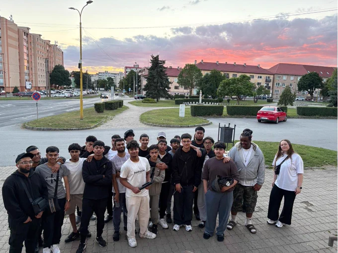 A group of young people gathered outdoors at sunset with a colorful sky. The mood is casual and social.