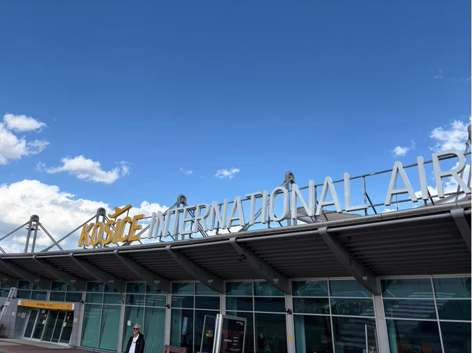 The entrance to Košice International Airport is captured under a clear blue sky. The signage prominently displays the airport's name, with a person standing outside.