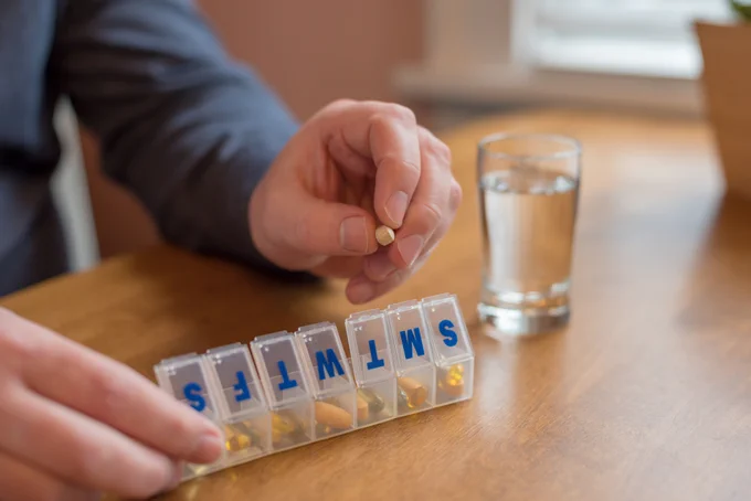 Person holding a small pill above a weekly pill organizer labeled with days and a glass of water nearby.