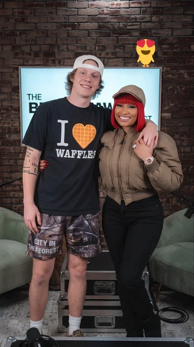 Nicki Minaj poses smiling with podcaster Bryce Crawford during a studio appearance. Bryce stands with his arm around Nicki, wearing a black “I ♥ WAFFLES” T-shirt, camouflage style shorts, white socks, and a backward white cap. Nicki wears a khaki bomber jacket, black fitted pants, a beige baseball cap, and a bright red bob hairstyle with blunt bangs. She accessorizes with a diamond ring, luxury watch, and long red nails. The pair stand in front of a brick wall and illuminated studio sign inside a podcast set, with lounge chairs, recording equipment, and production cases visible in the background. A small yellow bird with heart shaped eyes appears in the upper right corner, representing happybyrd.