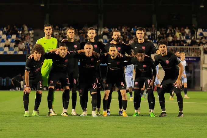 A soccer team poses for a group photo on the field before a match. The players are wearing black uniforms with a distinctive shield logo and are standing in two rows. The mood is focused and united.