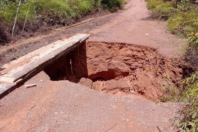 #DescriçãoDaImagem #Acessibilidade #ParaTodosVerem: Estrada de terra entre vegetação com um buraco profundo. Sobre o lado esquerdo da cratera, uma ponte de tábuas de madeira. Foto: Comunicação/MPF.