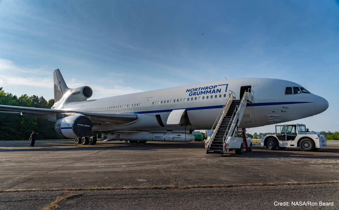 A large white airplane has a white rocket attached to its belly. A truck with a flight of stairs is pulled up to the forward boarding door, which is open. There is a band of blue running from the nose to the tail, just above the wings, and blue letters spell out “Northrop Grumman” to the left of the boarding door. A white tractor is hitched to the front of the airplane. The sky above is streaked lightly with white clouds. A person in a black shirt and pants stands under the wing. This image is watermarked with “Credit: NASA/Ron Beard.”