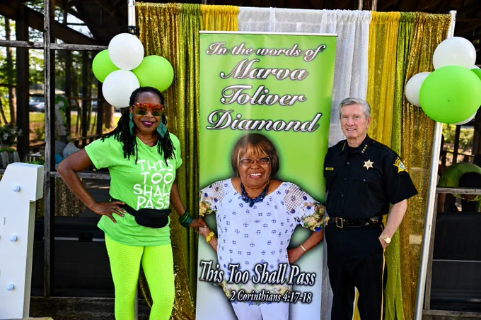 Two people stand in front of a banner featuring Marva Toliver Diamond and the phrase "This Too Shall Pass" with a Bible reference. The person on the left wears bright green clothing with text reading "This Too Shall Pass." The person on the right wears a sheriff's uniform. Green and white balloons are in the background.