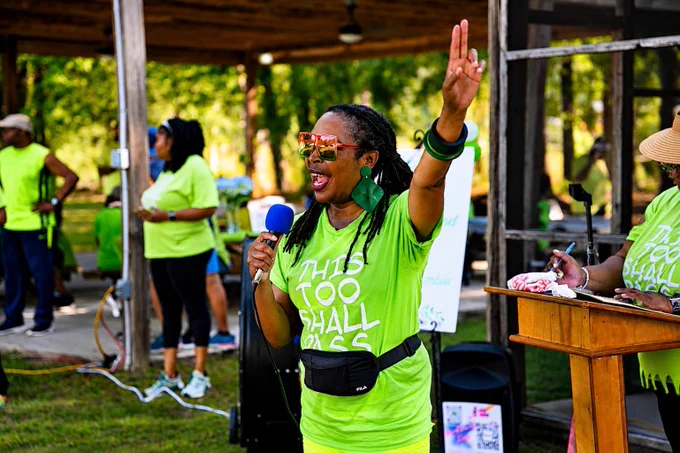 A person wearing a bright green shirt with "This Too Shall Pass" holds a microphone, raising a peace sign. They are speaking at an outdoor event near a podium. Others in matching green attire are in the background.