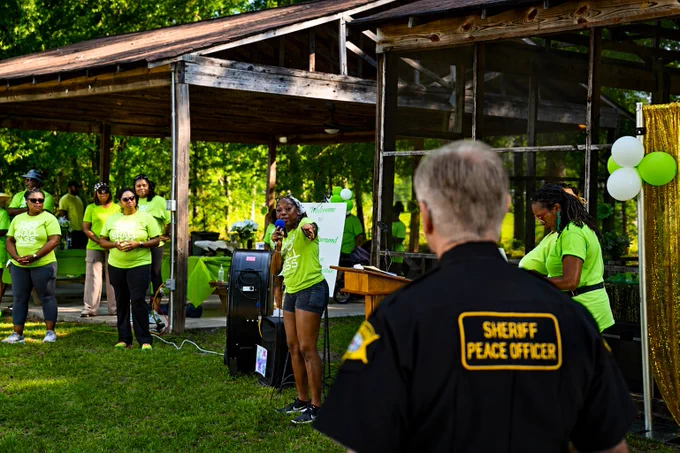 A person in a "Sheriff Peace Officer" shirt stands facing a group of individuals wearing bright green shirts in a park. One person is speaking into a microphone, while others stand nearby, surrounded by green and white decorations under a wooden pavilion.