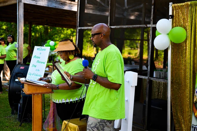 Two individuals in green shirts stand at a podium outdoors. One holds a microphone and a wooden plaque, while the other holds a large card. A sign on the podium reads "Welcome Metro Diamond Dog," with more text partially visible. White and green balloons decorate the area, and a gold curtain is in the background. Others are present in the background.