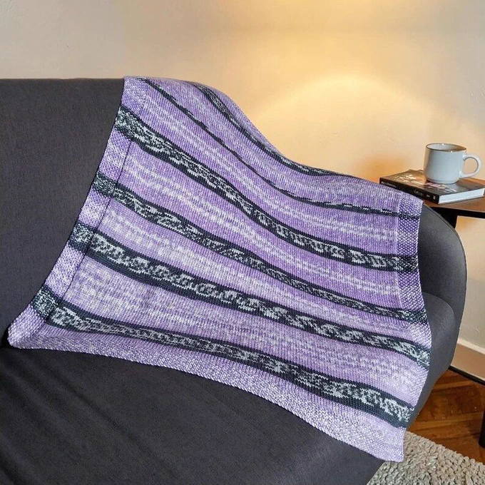 A lavender and dark grey striped hand-knitted baby blanket is draped over a dark grey sofa. In the background, a small side table holds a book and a white mug, softly lit by a warm lamp against a cream wall and wooden floor.