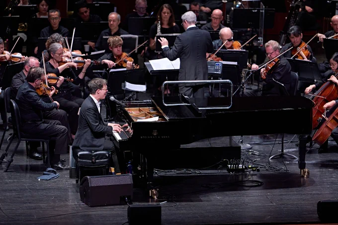 Ben Folds singing and playing the piano while Edwin Outwater conducts New Jersey Symphony musicians on stage.