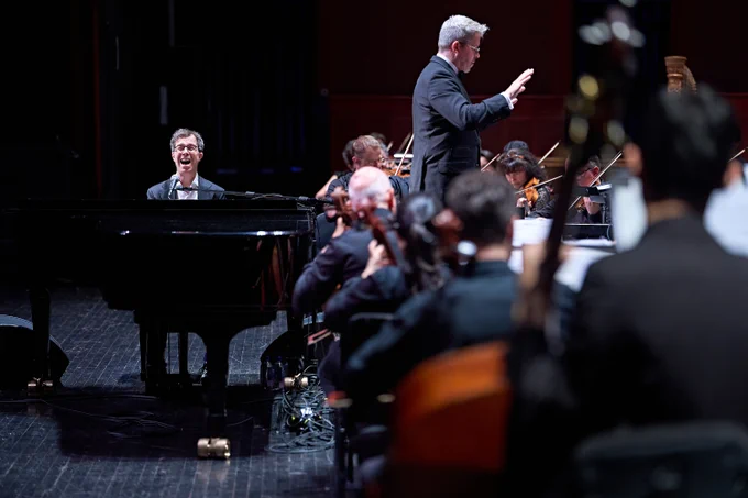 Ben Folds singing behind the piano while Edwin Outwater conducts New Jersey Symphony musicians on stage.