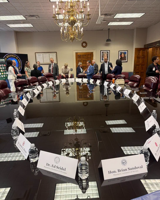 A large conference room with a reflective oval table is surrounded by red leather chairs. Nameplates for Dr. Ed Seidel and Hon. Brian Sandoval are visible on the table alongside water bottles. People are standing around the room, engaging in conversation. A chandelier hangs above the table, and framed documents and a round emblem are on the walls.