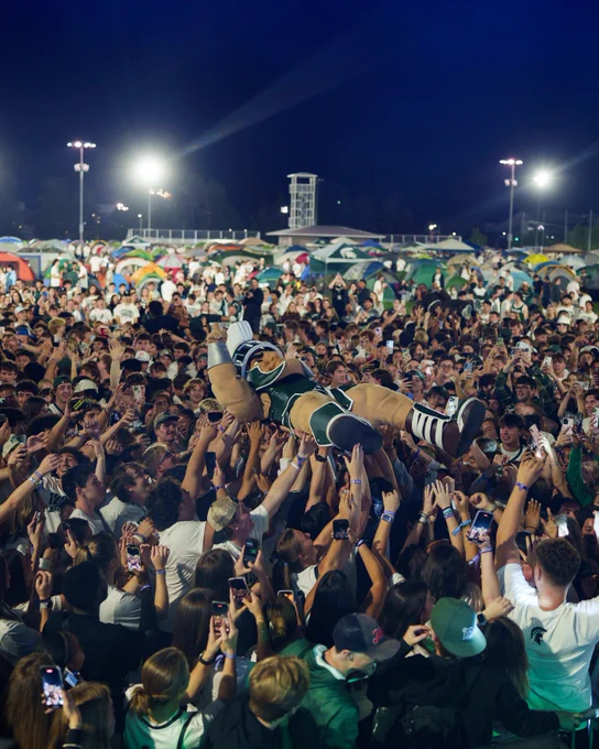 Sparty crowd surfs above a packed crowd during a nighttime campus event, surrounded by students, tents and raised phones.
