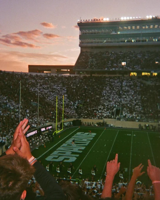 Fans cheer inside Spartan Stadium during a football game at sunset, with the crowd filling the stands and the field visible below.
