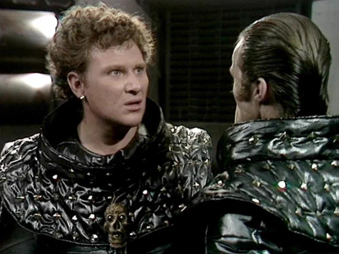 GPT 5.4 describes the image as: "A tense close-up from the classic BBC science-fiction series Blake’s 7, set in a stark, futuristic interior with low lighting and dark metallic surfaces. Two men face each other at close quarters, suggesting a confrontation or urgent exchange. On the left is Colin Baker, his expression alert and unsettled, wearing a glossy black, quilted costume with a high collar and a small skull-like ornament at the chest, adding to the dystopian, authoritarian feel of the setting. Opposite him is John J. Carney, seen mostly from behind and in profile, with slicked-back hair and a matching black, reflective outfit that reinforces the militaristic or institutional look. The scene’s costume design and claustrophobic framing evoke the show’s gritty, oppressive vision of the future."
The script search url for this tweet is
https://blake.torpidity.net/s/306/137