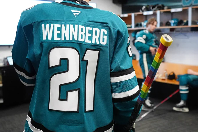 Picture of Alexander Wennberg, showing his back as he wears his teal Sharks jersey while he holds his stick with pride tape around the handle (in the Sharks dressing room)