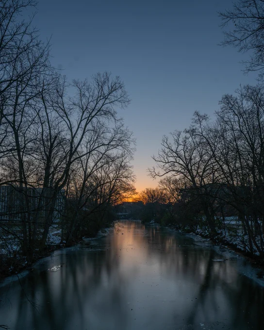 Frozen section of the Red Cedar River at sunset, reflecting bare trees and a blue-to-orange winter sky on Michigan State University's campus.