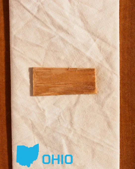 A rectangular piece of wood is placed on a white cloth. A light blue silhouette of the state of Ohio with the word "OHIO" beneath it is visible on the lower left corner of the image.