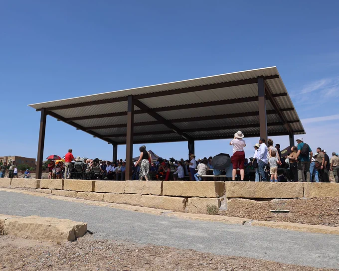 Large group of people gathered under a metal pavilion on a sunny day with clear blue sky.