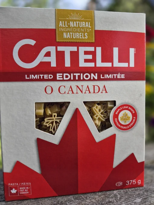 Picture of a box of Catelli brand limited edition "O CANADA" Maple leaf shaped pasta. It has the top part of the flag's Maple leaf in red at the bottom front of the box, with 2 plastic windows showing the pasta cut in Maple leaves.