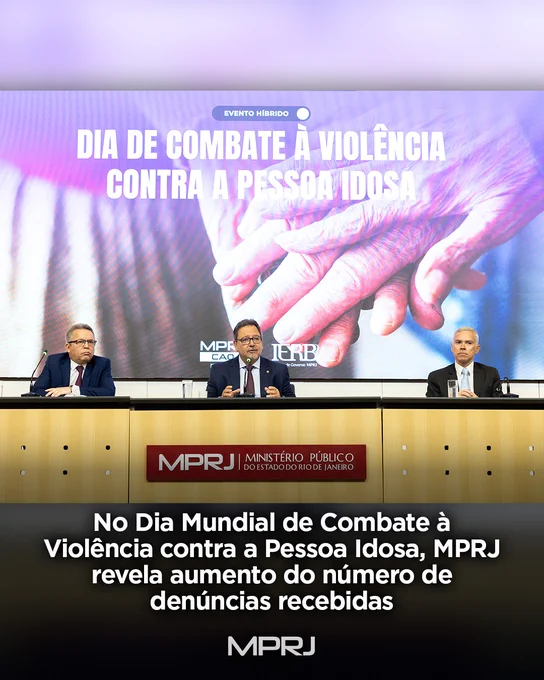Fotografia de uma mesa de evento institucional do Ministério Público do Estado do Rio de Janeiro (MPRJ). Três homens de terno estão sentados atrás da bancada, cada um com microfone e copo de água à frente. Ao fundo, um grande telão exibe a mensagem “Dia de Combate à Violência contra a Pessoa Idosa” sobre a imagem de mãos envelhecidas entrelaçadas. Na parte inferior da imagem, aparece o texto: “No Dia Mundial de Combate à Violência contra a Pessoa Idosa, MPRJ revela aumento do número de denúncias recebidas”. O logotipo do MPRJ está presente na bancada e na arte gráfica.