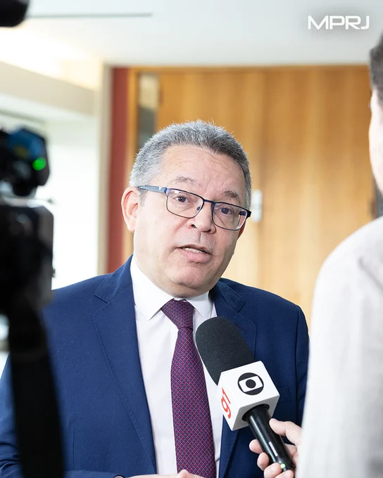 Fotografia em plano fechado do coordenador do Centro de Apoio Operacional das Promotorias de Justiça da Pessoa Idosa do MPRJ, promotor de Justiça Luiz Cláudio Carvalho de Almeida., vestido com terno azul, camisa branca e gravata vermelha, concedendo entrevista. Ele está voltado para um repórter que segura um microfone com a logomarca da TV Globo. À esquerda da imagem, parte de uma câmera de gravação aparece desfocada em primeiro plano. O ambiente é interno, com porta de madeira ao fundo e iluminação clara. O entrevistado mantém expressão séria enquanto responde às perguntas. O logotipo do MPRJ está no canto superior direito da imagem.