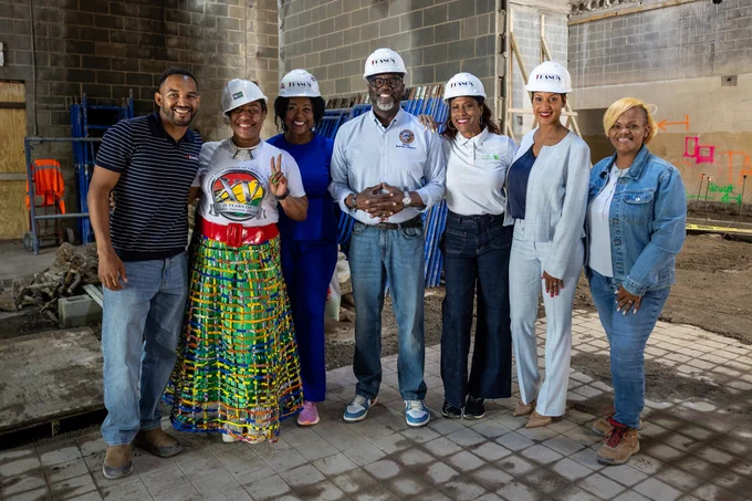 A group of seven individuals wearing hard hats stands together at a construction site. They are smiling and dressed in casual attire with some wearing jeans and others in colorful skirts or pants. The setting includes a partially built structure with exposed bricks and construction materials.