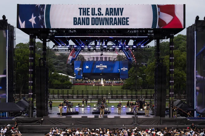 A large outdoor stage features a performance by The U.S. Army Band Downrange with musicians playing instruments. A prominent screen above the stage displays the group's name. In the background, the White House is visible. An audience is gathered in front of the stage.